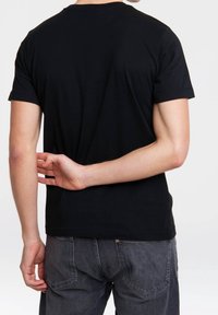 Osoba w czarnym T-shircie i ciemnoszarych dżinsach, z prawą ręką schowaną za plecami, krzyżując palce.