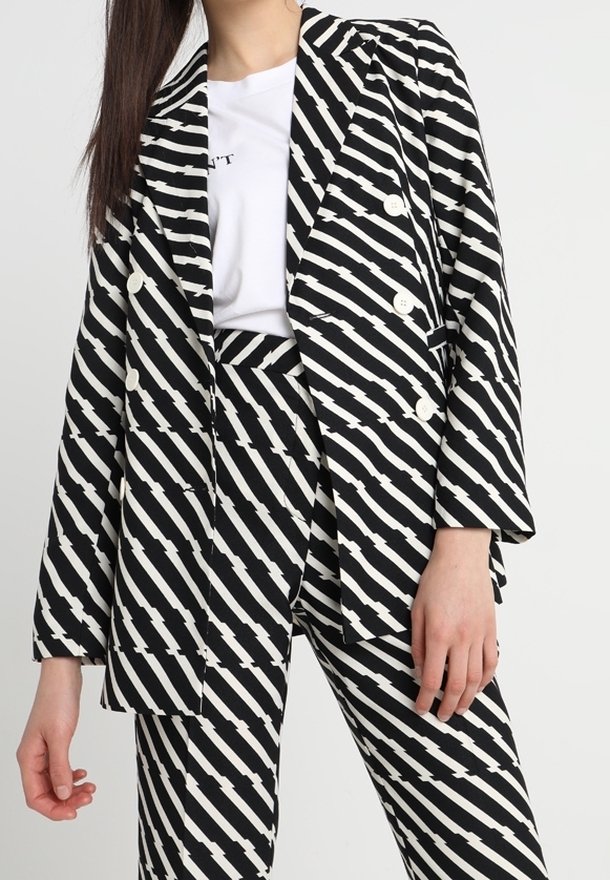 Femme portant un blazer croisé à rayures en zigzag noir et blanc et un pantalon assorti avec une chemise blanche en dessous.