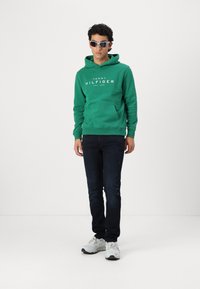 Hanorac verde din bumbac, cu buzunar frontal tip kangaroo și logo-ul alb "TOMMY HILFIGER", asortat cu blugi închisi la culoare și pantofi sport albi.