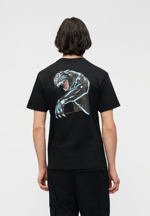 Persona con cabello oscuro de longitud media que lleva una camiseta negra con una ilustración de una pantera negra rugiente en la espalda, sobre un fondo liso.