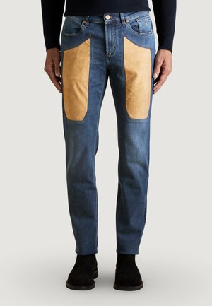 Jeans blu a gamba dritta con grandi toppe in camoscio color sabbia sulla parte anteriore delle cosce, indossati con stivali neri e una maglia scura a maniche lunghe.