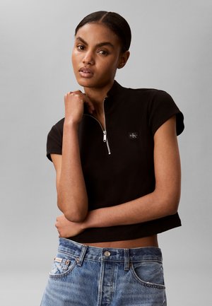 Kvinde poserende med en hånd nær hagen, iført en sort kortærmet top med lynlås og blå cowboystøvler mod en grå baggrund.