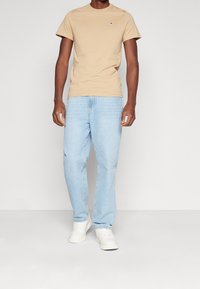 Světle hnědé bavlněné tričko s krátkým rukávem, kombinované se světle modrými džínami. Model nosí bílé tenisky a stojí na neutrálním pozadí.