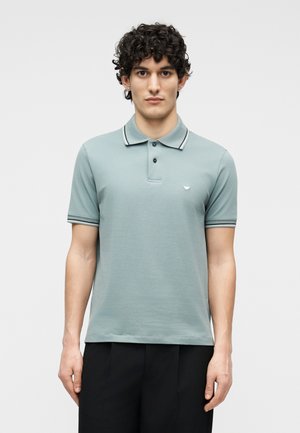 Νεαρός με κατσαρά μαλλιά που φοράει ανοιχτό πράσινο πόλο με κοντά μανίκια και μαύρη επένδυση στο γιακά, καθώς και μαύρο παντελόνι, στέκεται με το σώμα στραμμένο προς τα εμπρός.