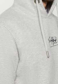 Homme portant un sweat à capuche gris clair avec cordons à l'avant et le logo noir brodé "Squidclub EST. 2017" sur la poitrine.