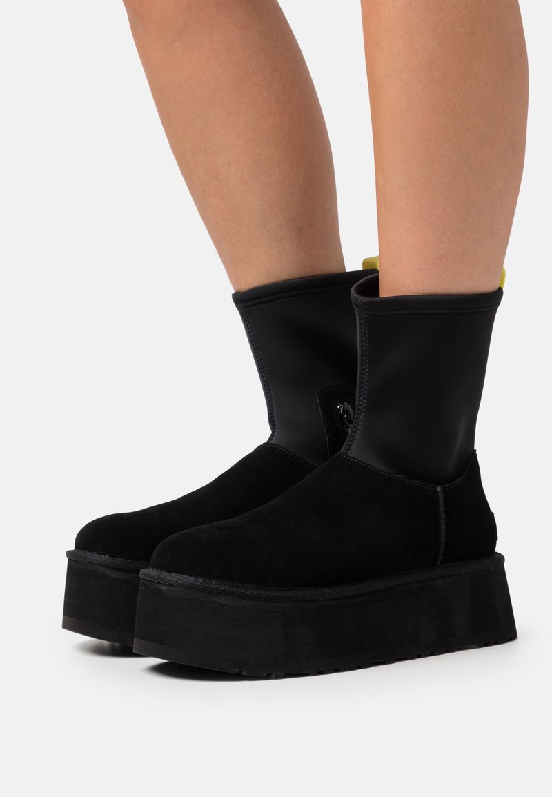 Bottines noires à plateforme avec semelles épaisses, portées par des jambes nues sur un fond clair uni.