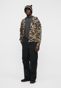 Mees triibulise kootud mütsi, kamuflaažiga lukuga jope, tumedate pükste ja mustade kingadega, seisab käed taskutes.