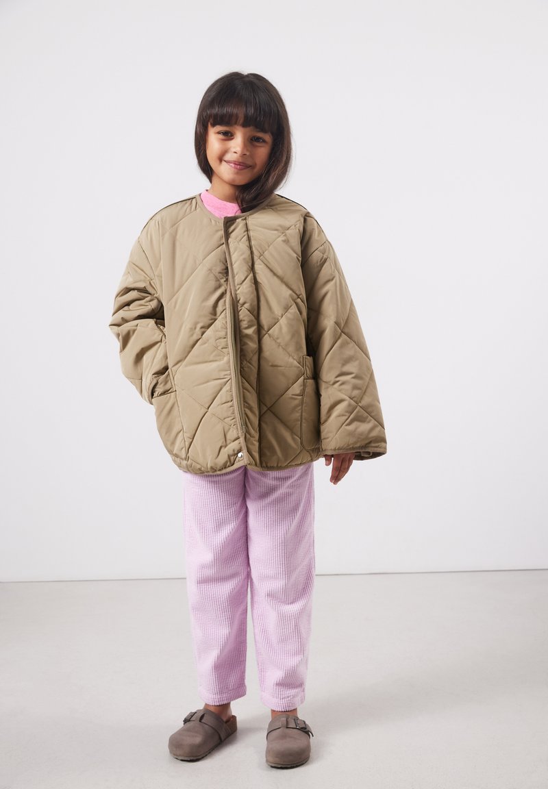 Jeune fille souriante, portant une veste matelassée beige surdimensionnée, un pantalon en velours côtelé rose et des chaussures marron faciles à enfiler, debout sur un fond uni.