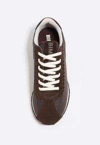 Zapatilla marrón con cordones blancos, detalle de lengüeta de ante con bordes en forma de ola y cuello acolchado blanco, vista desde arriba sobre un fondo blanco.
