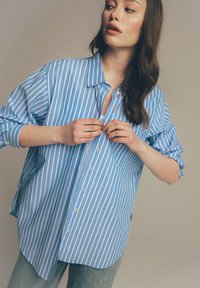 Jasnoniebieska, w paski koszula z guzikiem o luźnym kroju, z kołnierzykiem i długimi rękawami, prezentowana na modelu.