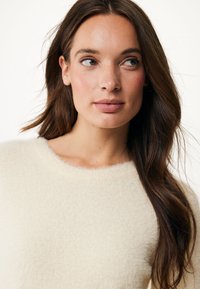 Pull crème, moelleux, à col rond avec une texture douce et des détails fins. Modèle avec des cheveux longs montré de l'épaule vers le haut.