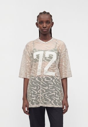 Personne noire aux cheveux tressés portant un T-shirt en maille beige ample avec le numéro 12 sur un haut à motifs et un pantalon noir, regardant vers l'avant.
