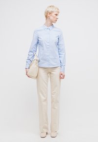 Άτομο με κοντά ξανθά μαλλιά φοράει ένα ανοιχτό μπλε πουκάμισο με κουμπιά, κρεμ παντελόνι και κρατά μια κρεμ τσάντα, κοιτάζοντας προς τα δεξιά.