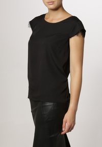 Femme portant un haut noir à manches courtes avec bordure en dentelle et une jupe en cuir noir, debout devant un fond clair uni.