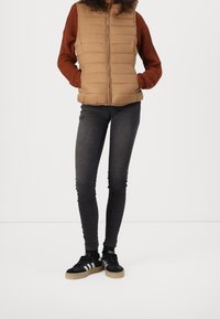 Braun gepolsterte Weste mit hohem Kragen und Reißverschluss, kombiniert mit einem rostfarbenen Strickpullover, schwarzen schmalen Jeans und schwarzen Sneakers mit weißen Streifen.