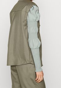 Oliwkowa kamizelka na jasnoniebieskiej, marszczonej koszuli z długim rękawem. Koszula ma marszczony materiał przy nadgarstkach i luźny fason na ramionach.