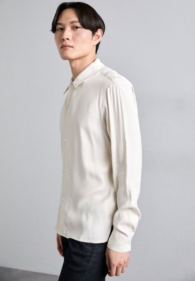 Licht beige lange mouwen shirt met een kraag, knoopsluiting aan de voorkant en subtiele plooien op de schouders. Draagt met donkere jeans.
