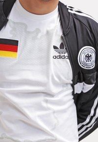 Maglietta sportiva bianca con texture a pois, logo Adidas nero, indossata sotto una giacca a vento nera con tre strisce bianche e toppa della bandiera tedesca.