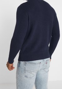 Granatowy sweter prążkowany z wysokim kołnierzem, długimi rękawami i teksturowanym wykończeniem, zestawiony z jasnoniebieskimi dżinsami.