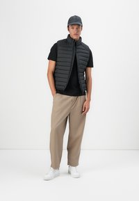 Sort puffer vest med høj krave, parret med en sort t-shirt og beige bukser. Hvide sneakers fuldender outfittet.