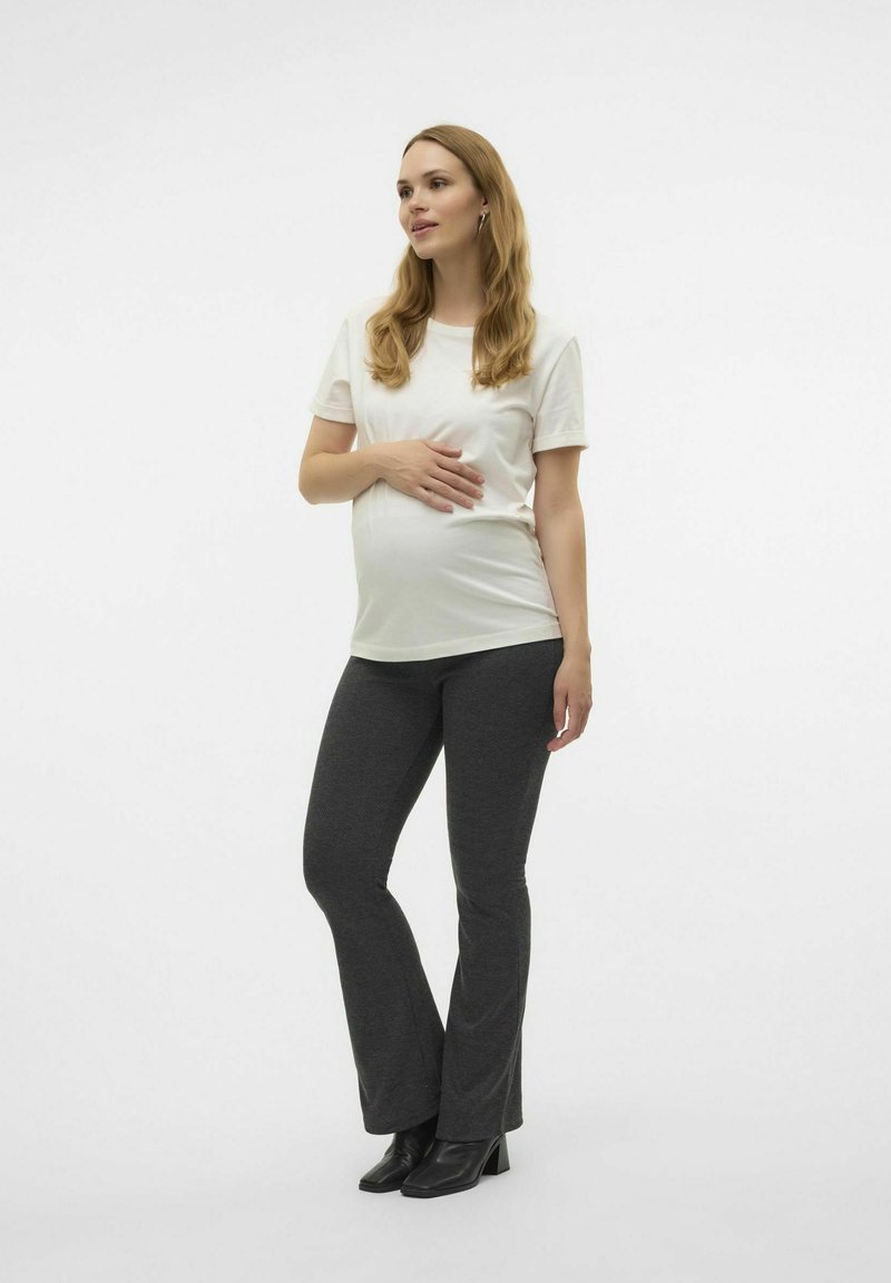 Graviditets t-shirt i mjukt vitt tyg, ihop med mörkgrå flare leggings. Modellen poserar med handen på magen, iförd svarta klackstövlar.