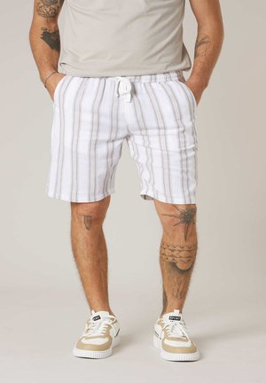 Mann trägt weiß-grau gestreifte Shorts mit Kordelzug, beige-weiße Sneaker und ein helles Hemd, zeigt Tätowierungen an Armen und Beinen.