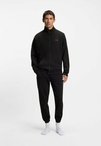 Completo tuta nera realizzato in tessuto texturizzato, composto da giacca con zip e colletto alto con logo, abbinato a pantaloni affusolati e calzini bianchi.