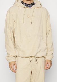 Sudadera con capucha de corduroy beige, con un bolsillo frontal y cordones en el cuello. Tejido texturizado con detalles acanalados y puños elásticos.