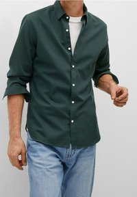 Dark green button-up shirt with a collared neck, long sleeves rolled to the elbows, paired with a white t-shirt and blue jeans.