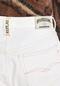 Pantalones vaqueros de mezclilla blanca con una etiqueta de marca amarilla y azul, con costuras naranjas y un bolsillo trasero con un diseño estampado.