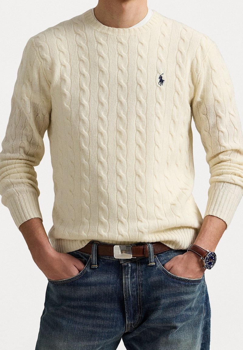 Homme portant un pull crème à torsades avec logo, ceinture marron, jean bleu et montre bleu marine, mains dans les poches, sur fond blanc.