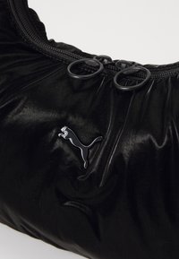 Sac rembourré noir avec une texture brillante, équipé de deux tirettes de fermeture éclair et d'un logo Puma noir bien en vue. Forme compacte et douce.