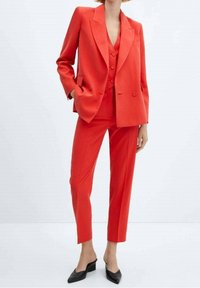 Costume ajusté rouge composé d'un blazer croisé et d'un pantalon slim, en tissu lisse, associé à des escarpins noirs pointus.