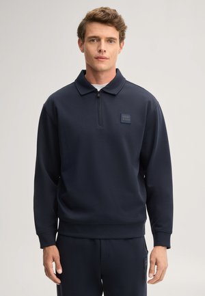 Junger Mann trägt einen marineblauen Viertel-Zip-Sweatshirt mit kleinem Logoaufnäher auf der linken Brust, steht vor einem schlichten hellen Hintergrund.