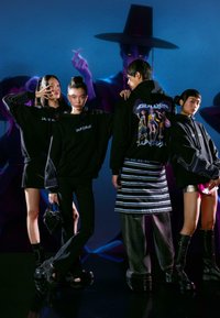 Štyria jednotlivci majú na sebe čierne mikiny s grafickými motívmi, doplnené rôznymi textúrami a vzormi. Pozadie tvorí farebné digitálne umenie.