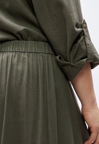 Primer plano de una persona vistiendo un vestido verde oliva con cintura elástica fruncida y manga remangada asegurada con una presilla de botón.