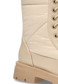 Botín acolchado en color beige con punta redonda, diseño de cordones, tela texturizada y suela de goma gruesa. Incluye costuras decorativas.