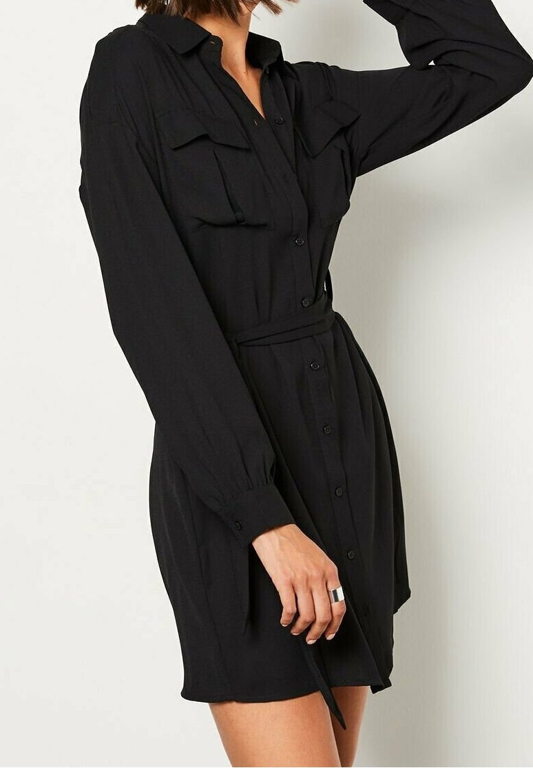 Femme portant une robe noire à manches longues boutonnée, avec des poches poitrine et une ceinture à la taille, tenant son bras au-dessus de la tête.