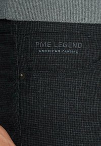 Ciemne spodnie teksturowane z plecionym wzorem, z czarnym guzikiem i metką z napisem "PME LEGEND AMERICAN CLASSIC" w kolorze niebieskim.