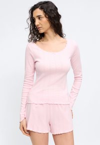 Γυναίκα με σγουρά σκούρα μαλλιά, φορώντας ροζ ανοιχτό ριχτό μακρυμάνικο τοπ και αντίστοιχα σορτς με διακριτά υφή patterns.