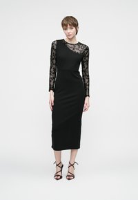 Naine kannab musta midi kleiti, millel on üks läbipaistev helmestega varrukas ja vastav rihmadega kõrged kontsad, lausal valgel taustal.