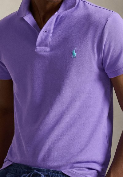 Mees kannab sobivat lavendlivärvi polo särki, millel on vasakul rinnal väike türkiissinine tikitud logo, kombineeritud tumedate pükstega.