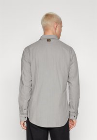 Grå långärmad skjorta med knapp-ner krage, subtil struktur, rundad nederkant, och en etikett vid nacke. Svarta knappar på ärmsluten.