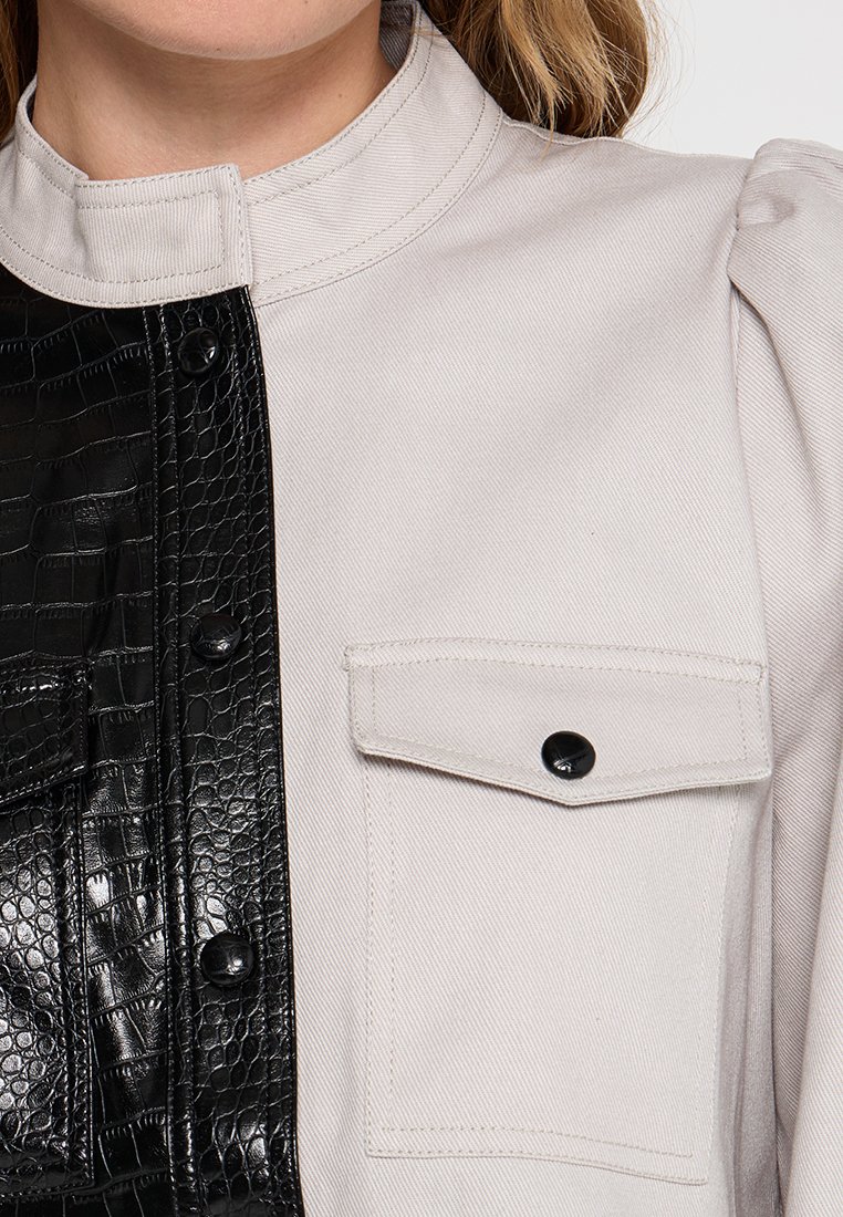 Jasnoszara kurtka z czarnymi detalami z materiału przypominającego skórę krokodyla. Posiada stójkę, zapięcie na zatrzask oraz jedną kieszeń na piersi po lewej stronie.