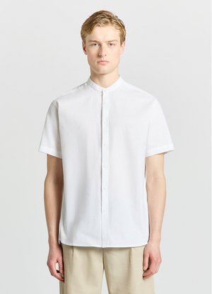 Jeune homme aux cheveux blonds courts portant une chemise blanche à manches courtes avec col mandarin et un pantalon beige, debout devant un fond clair uni.