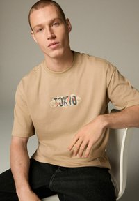 Camiseta de manga corta beige con gráfico "TOKYO" en azul y rojo. Combinada con jeans oscuros, sentado en una silla blanca frente a un fondo neutro.