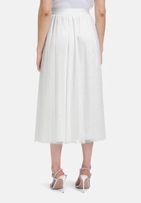 Jupe en tulle blanche avec des plis, longueur mi-mollet et ceinture en satin. Associée à des sandales à talons argentées, dévoilant une texture légère et aérienne.