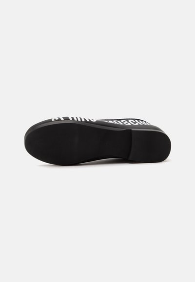 Chaussure plate noire vue de la semelle avec une semelle extérieure lisse et un petit talon, une partie du texte blanc visible sur le bord supérieur.