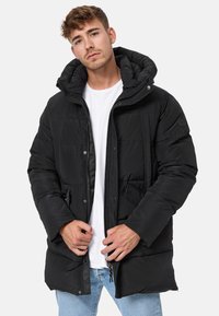 Chaqueta acolchada negra con capucha, con cierre de cremallera en la parte delantera, botones a presión, textura acolchada y bolsillos laterales. Llevada sobre una camiseta blanca.