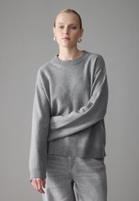 Grauer Pullover mit geripptem Halsausschnitt und Bündchen, aus weicher Wolle gefertigt, hat einen lockeren Schnitt und lange Ärmel, kombiniert mit hellgrauen Jeans.
